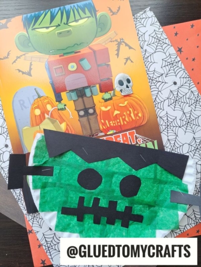 A green paper plate Frankenstein craft sits atop a Halloween-themed book decorated with cartoon Frankenstein, pumpkins, skulls, and spider webs. The handle @GLUEDTOMYCRAFTS is displayed at the bottom.