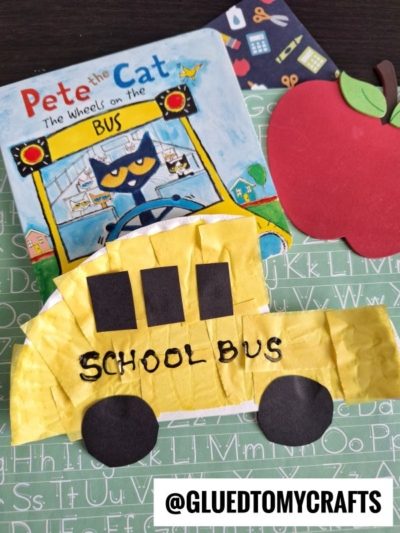 A children’s craft scene featuring the book “Pete the Cat: The Wheels on the Bus,” a cheerful paper school bus made from tissue and paper, and a red paper apple, all on an alphabet background. Text: @GLUEDTOMYCRAFTS.