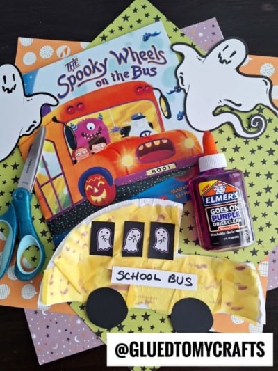 A Halloween-themed craft setup with scissors, glue, papers, a book called *The Spooky Wheels on the Bus*, ghost cutouts, and a yellow paper plate bus decorated with ghost faces—perfect for creating your own school bus craft or spooky paper school bus.