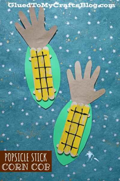 Two handmade craft stick corn cobs made from yellow popsicle sticks, green and brown paper, and black marker are arranged on a blue background with white star patterns. Text reads Popsicle Stick Corn Cob.