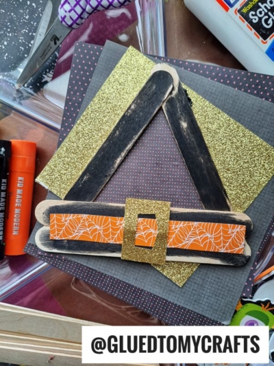 A popsicle stick witch hat craft made from black and gold glitter paper, wooden craft sticks, and orange spider web ribbon, surrounded by glue sticks and craft supplies.