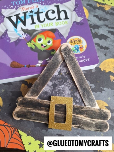 A charming popsicle stick witch hat with a gold buckle sits on a table near the book There’s a Witch in Your Book. The table features Halloween-themed designs, perfectly complementing the playful popsicle stick witch hats on display. Text reads @GLUEDTOMYCRAFTS.