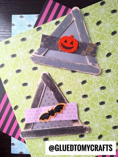 Two triangle popsicle stick witch hats, each decorated with Halloween-themed felt shapes, sit on layered green spiderweb and striped papers. The bottom right corner has the text @GLUEDTOMYCRAFTS.