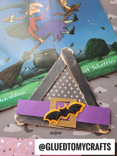 A triangle-shaped craft, resembling popsicle stick witch hats, is made from black-painted sticks, decorated with a purple strip and a black-and-yellow bat, and displayed near a Room on the Broom book cover featuring a witch on a broomstick.