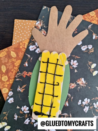 A fall-themed craft featuring a handprint cutout as a corn husk, yellow painted craft sticks or popsicle sticks as corn kernels, and black yarn as corn lines—all arranged on floral paper. This adorable popsicle stick corn project is complete with the @GLUEDTOMYCRAFTS watermark at the bottom.