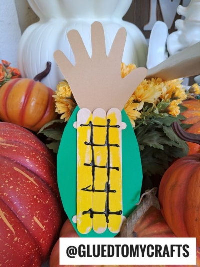A hand-shaped paper cutout is decorated as an ear of corn with green and yellow paper, black lines for kernels, and surrounded by pumpkins and flowers—perfect to pair with a popsicle stick corn craft. The text @GLUEDTOMYCRAFTS is at the bottom.
