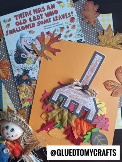 Children’s book *There Was an Old Lady Who Swallowed Some Leaves!* paired with a raking leaves kid craft featuring a paper rake and faux leaves, all set among autumn decorations and a small scarecrow doll.