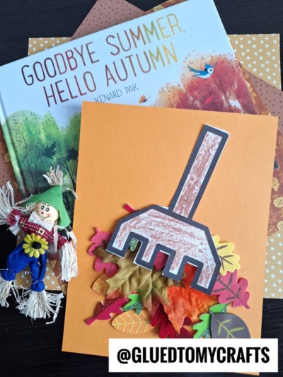 A children’s book titled Goodbye Summer, Hello Autumn, autumn-themed papers, a small scarecrow figure, and a raking leaves kid craft made from a paper rake with artificial leaves are arranged on a table. @GLUETOMYCRAFTS is written at the bottom.