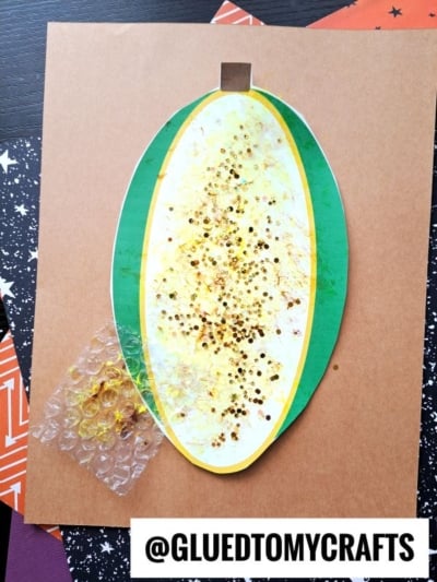 An oval-shaped craft with yellow and green layers, gold glitter, and a piece of bubble wrap on brown paper resembles bubble wrap corn on the cob. The username @GLUEDTOMYCRAFTS appears at the bottom.
