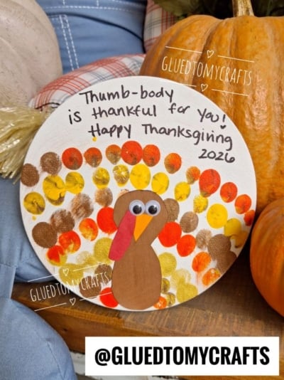 A delightful Thanksgiving turkey kid craft featuring painted thumbprints for feathers, googly eyes, and a beak on a round canvas. The text reads, Thumb-body is thankful for you! Happy Thanksgiving 2026.