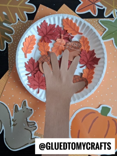 A paper plate fall tree displays colorful paper leaves and a brown paper hand, surrounded by autumn-themed cutouts like a squirrel and pumpkin. The username @GLUEDTOMYCRAFTS is visible at the bottom.