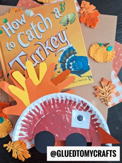A children’s book titled How to Catch a Turkey sits on a table surrounded by autumn leaves, pumpkins, and an orange-and-red turkey kid crafts project made from paper plates and handprints. A sticker reads @GLUEDTOMYCRAFTS.