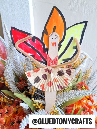A handmade turkey kid craft with colorful leaf wings, a smiling face, and a skirt made from decorated cupcake liners, placed among orange flowers. @GLUEDTOMYCRAFTS is written at the bottom.