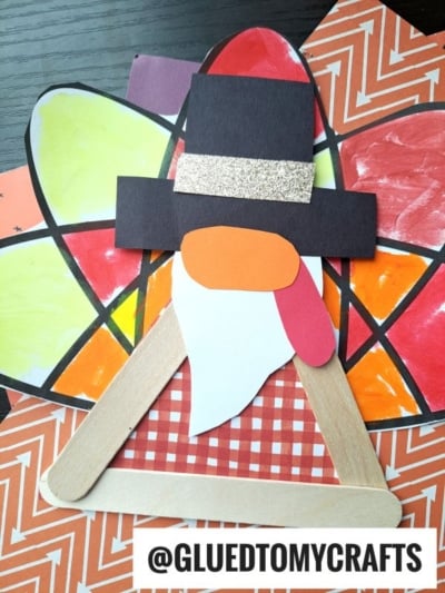 A Thanksgiving craft perfect for turkey kid crafts: a pilgrim face with a black hat, white beard, and orange details, made from colored paper and craft sticks. The background features colorful, painted paper feathers.
