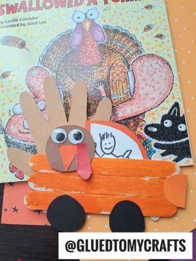 A handmade turkey kid craft made with painted popsicle sticks and paper sits atop a children’s book featuring a turkey illustration. The handle @GLUEDTOMYCRAFTS is displayed at the bottom.