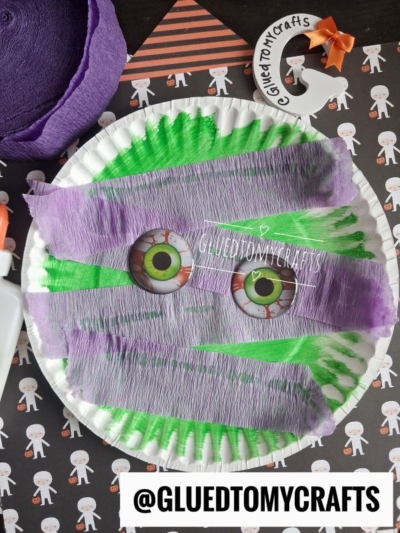 A paper plate mummy kid craft featuring green and purple crepe paper strips, large cartoon eyes, and a @gluedtomycrafts handle, set on a Halloween-themed background.
