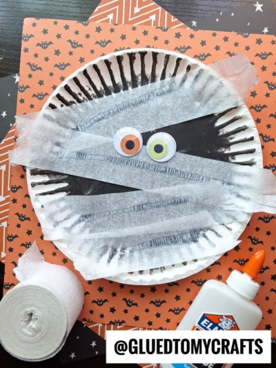 A fun paper plate mummy kid craft featuring large googly eyes, white gauze strips, a bottle of glue, and craft paper with bat and star patterns in the background.