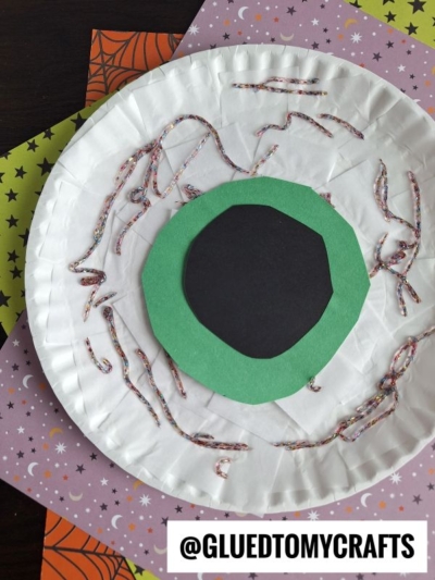 A spooky paper plate eyeball craft features a bold black center on vibrant green paper, accented with glitter glue veins for a realistic touch. Colorful Halloween-themed paper adds festive flair in the background, making this clever paper plate eyeball project perfect for holiday crafting fun. @GLUEDTOMYCRAFTS is labeled at the bottom.
