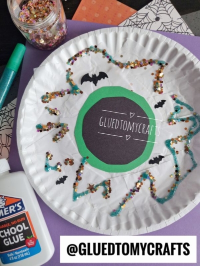 A paper plate eyeball decorated as a spooky masterpiece with glitter, sequins, bats, school glue, and @GLUEDTOMYCRAFTS written in the center. Craft supplies and patterned paper surround the plate.