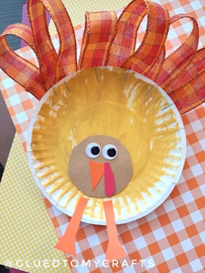 A charming Paper Plate & Ribbon Turkey craft featuring an orange and yellow painted plate, festive ribbon tail feathers, a brown paper face, googly eyes, and playful orange legs, all arranged on an orange checkered background.