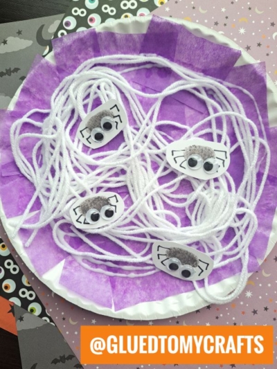 A paper plate spider web with purple tissue, white yarn woven across the center, and four playful paper spiders featuring googly eyes and drawn legs. Halloween-themed papers surround the plate, creating a festive display. @GLUEDTOMYCRAFTS is highlighted in an orange banner.