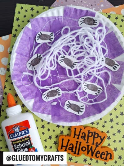 A Halloween craft featuring a paper plate spider web made with purple tissue, white yarn arranged like a web, black spider cutouts, a bottle of Elmer's glue, and a Happy Halloween sign on a festive background.
