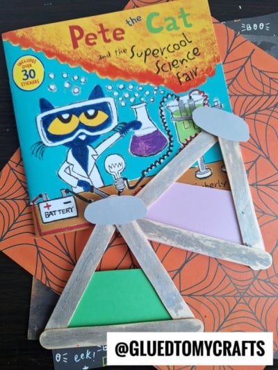 A “Pete the Cat” science fair book rests on a Halloween-themed orange and black surface, topped with a handmade Popsicle Stick Beaker balance scale craft. @GLUEDTOMYCRAFTS is in the bottom right corner.