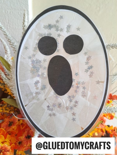 A ghost face made with tissue paper, star-shaped glitter, and black cut-outs for the eyes and mouth—reminiscent of playful Stained Glass Ghosts. The craft is displayed among orange flowers, with @GLUEDTOMYCRAFTS label at the bottom.