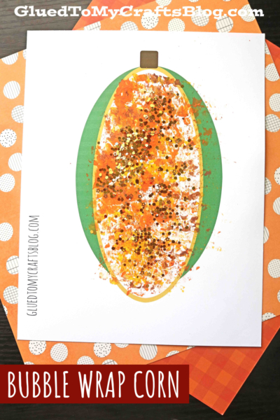 A children's craft features bubble wrap stamped corn on the cob, decorated with orange, yellow, and brown paint plus glitter, on a white sheet with a green husk outline. The paper rests on orange, polka-dotted sheets.
