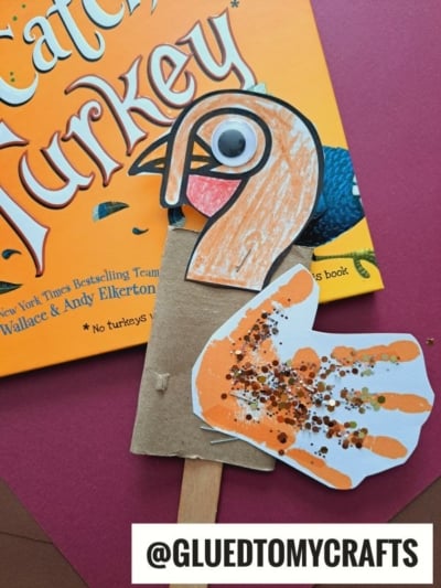 A handmade turkey kid crafts project made from a colored paper handprint, cardboard, googly eye, and popsicle stick sits on an orange “Catch a Turkey” book cover. The craft sparkles with glitter. Text: @GLUEDTOMYCRAFTS.