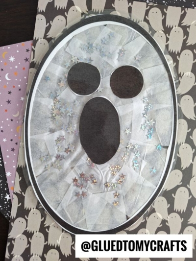 A ghost face craft made with white tissue paper, an oval outline, cut-out eyes and mouth, and star-shaped sequins resembles stained glass ghosts. Displayed on patterned paper with ghost and star designs. Text reads @GLUEDTOMYCRAFTS.