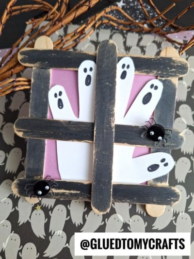 Three paper ghosts peek out from behind a popsicle stick ghost window, crafted from black wooden craft sticks, with two small fake spiders attached. Halloween-themed paper with ghost prints is in the background. @GLUEDTOMYCRAFTS is at the bottom.
