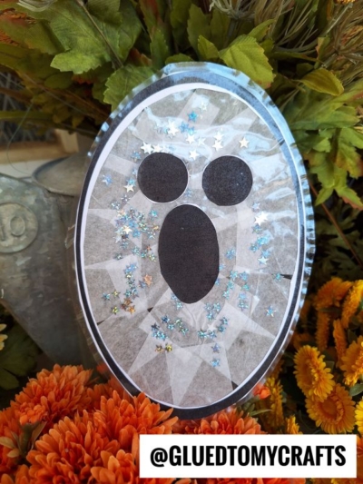 A handmade Stained Glass Ghosts craft with black oval eyes and mouth, decorated with star-shaped glitter, is propped among orange flowers. The Instagram handle @GLUEDTOMYCRAFTS is displayed at the bottom.