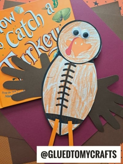 A handprint turkey kid craft made of colored paper and googly eyes sits atop the book How to Catch a Turkey. The turkey has a brown body, orange beak, and two handprint wings. @GLUEDTOMYCRAFTS is written below.