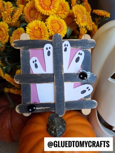 A Popsicle Stick Ghost Window Halloween craft features handprint ghosts on pink paper behind black craft stick bars, with plastic spiders attached. Yellow flowers and a pumpkin complete the scene. Text reads @GLUEDTOMYCRAFTS.