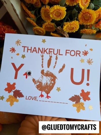 A card with fall leaves and stars reads “Thankful for You!” The “O” in “You” is a googly eye, and a handprint forms a turkey—perfect for turkey kid crafts. Orange flowers surround the card. Text at the bottom: “@GLUEDTOMYCRAFTS.”.