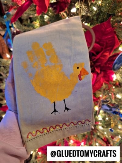 A hand holds a towel with a yellow handprint turkey kid craft, featuring painted details and a wavy trim, in front of a decorated Christmas tree. @GLUEDTOMYCRAFTS is written at the bottom.