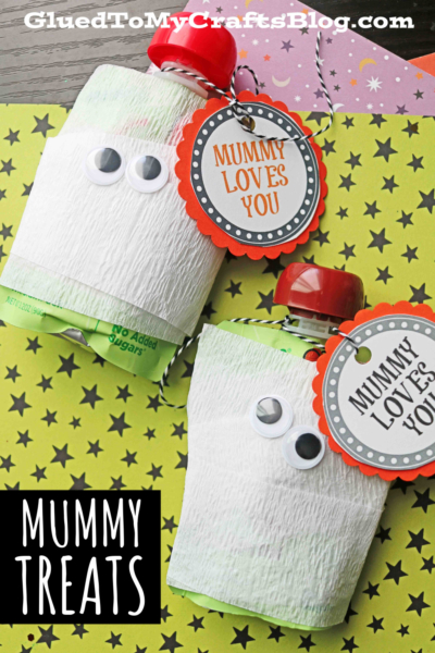 Two mummy squeeze snacks—applesauce pouches wrapped in white crepe paper with googly eyes and “Mummy Loves You” tags—rest on yellow paper with black stars. Text reads “Mummy Treats.”.