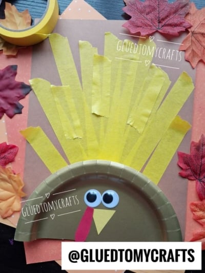 A fun turkey kid craft made with a paper plate, yellow paper strip feathers, googly eyes, a red and yellow beak, and artificial leaves. @GLUEDTOMYCRAFTS is written on the image.