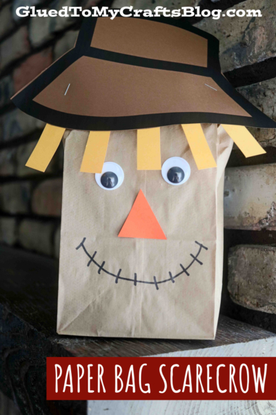 A Paper Bag Scarecrow craft featuring googly eyes, a paper hat, yellow paper hair, an orange triangle nose, and a stitched smile. Text reads GluedToMyCraftsBlog.com and PAPER BAG SCARECROW.