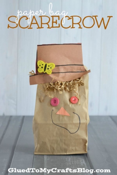 A charming Paper Bag Scarecrow with a brown hat, button eyes, a paper nose, and a marker-drawn smile is adorned with a yellow paper butterfly, all displayed on a gray wooden surface. Text reads Paper Bag SCARECROW.