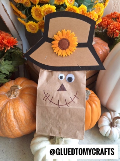 A charming Paper Bag Scarecrow puppet with googly eyes, a drawn stitched smile, and a paper hat decorated with a sunflower sits among orange and white pumpkins and autumn flowers. The text @GLUEDTOMYCRAFTS is at the bottom.