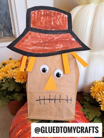 This adorable Paper Bag Scarecrow craft features a red paper hat, yellow paper hair, large googly eyes, an orange triangle nose, and a stitched mouth. It's surrounded by pumpkins and yellow flowers for a festive autumn touch.