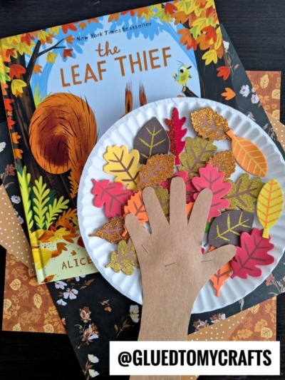 A paper plate fall tree with colorful paper leaves and a hand cutout sits on top of the book The Leaf Thief by Alice Hemming. Craft materials and autumn-themed paper are in the background.