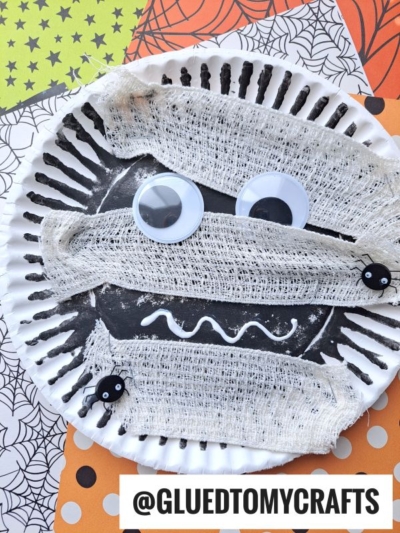 A spooky paper plate mummy kid craft featuring googly eyes, white gauze, black painted lines, and tiny spider decorations, all displayed on a colorful Halloween-themed background. Text reads @GLUEDTOMYCRAFTS.
