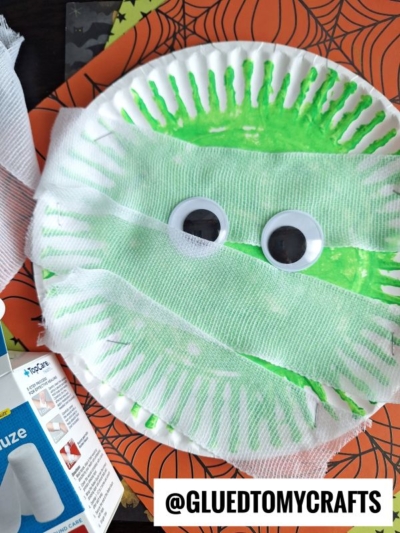A paper plate mummy kid craft, painted green with big googly eyes and gauze strips, sits on a Halloween-themed spiderweb background. @GLUEDTOMYCRAFTS text is visible.