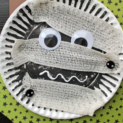 A fun mummy kid craft: a paper plate mummy decorated with black paint, giant googly eyes, gauze strips, and small plastic spiders on green star-patterned and purple paper. Text reads PAPER PLATE MUMMY.