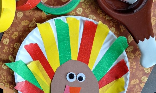 A colorful turkey craft made from a paper plate, colored tissue strips, googly eyes, and brown paper sits on fall-themed paper—perfect inspiration for turkey kid crafts. Tape rolls and a brown turkey-shaped bowl are nearby. Text reads @GLUEDTOMYCRAFTS.