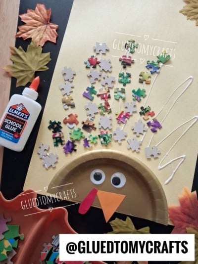 A fun turkey kid craft using a paper plate, puzzle piece feathers, googly eyes, colored paper beak and wattle—plus Elmers glue, leaves, and a tray nearby. @GLUEDTOMYCRAFTS is on the image.