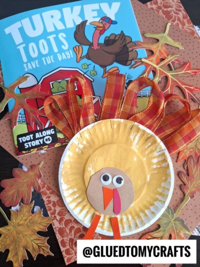 A childrens book titled Turkey Toots Save the Day! is displayed with a Paper Plate & Ribbon Turkey craft, autumn leaves, and a @GLUEDTOMYCRAFTS label. The turkey features colorful ribbon feathers and a simple face.